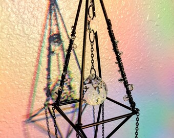 May include: A black metal geometric frame with hanging crystal prisms. The frame is in the shape of a pyramid with a square base. The crystals are clear and reflect light.