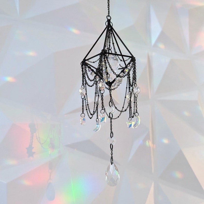 May include: A black metal chandelier with crystal drops and star-shaped accents hangs from a white ceiling. The chandelier casts rainbows on the white wall behind it.