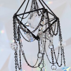 May include: A black metal geometric frame with a crystal sphere hanging from it. The frame is adorned with chains and crystals, creating a cascading effect. The crystals are iridescent and sparkle in the light.
