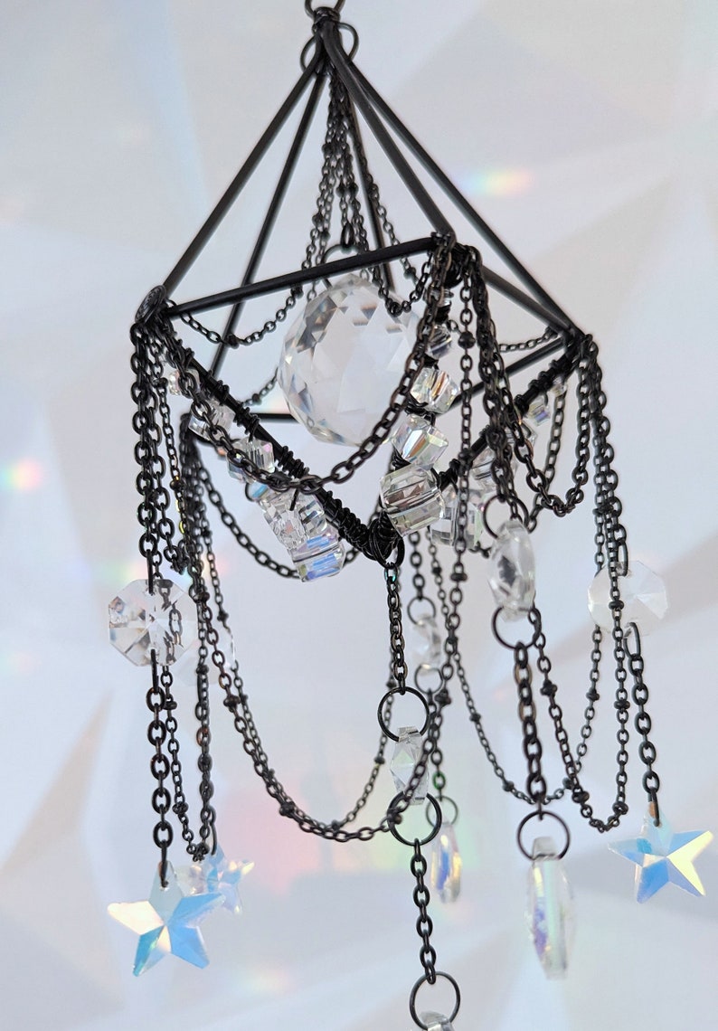 May include: A black metal geometric frame with a crystal sphere hanging from it. The frame is adorned with chains and crystals, creating a cascading effect. The crystals are iridescent and sparkle in the light.