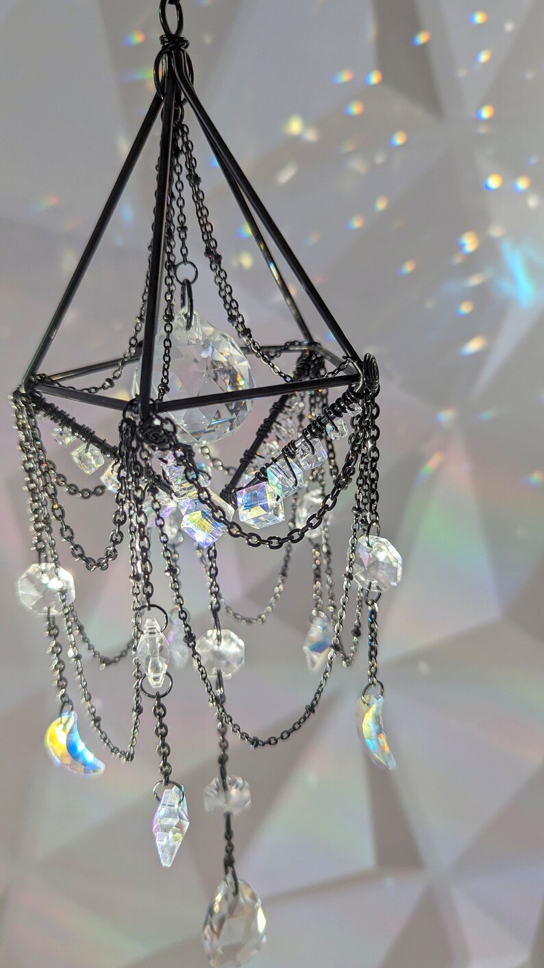 May include: A black metal geometric chandelier with crystal prisms and a large crystal orb hanging from the center. The chandelier is decorated with chains and has a modern design.