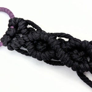 May include: Close-up of a macrame plant hanger in black. The hanger features a purple ring at the top and knotted black rope forming a decorative pattern. The design includes multiple strands of black rope extending from the knotted section.