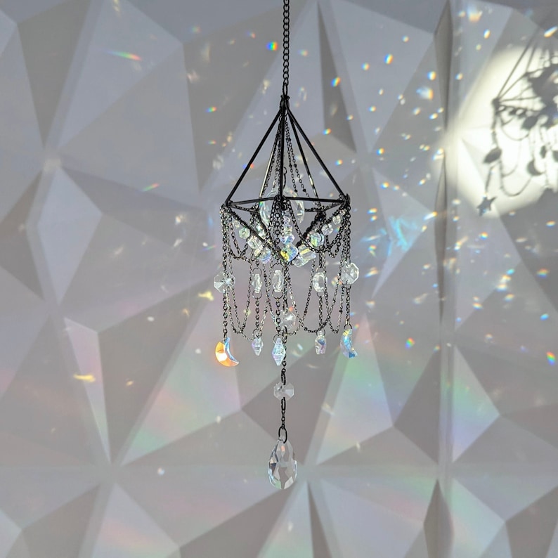 May include: A black metal chandelier with crystal prisms hanging from a white ceiling. The chandelier is in the shape of a pyramid with a pointed top and a square base. The crystals are arranged in a symmetrical pattern around the chandelier.