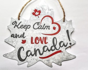 Op de afbeelding: Een wit houten bord in de vorm van een esdoornblad met de tekst "Keep Calm and Love Canada!" in zwarte en rode letters. Een rood hart met een klein esdoornblad erin bevindt zich boven het woord "Love".