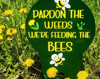 May include: A green metal sign with the text "Pardon the weeds we're feeding the bees" in yellow lettering. The sign has two cartoon bees with white bodies and yellow stripes. The sign is in a field of green grass and yellow dandelions.