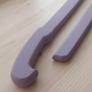 May include: Two smooth, matte lavender-colored hockey sticks featuring a curved handle design. The sticks have a uniform, solid light purple finish with rounded edges and a sleek, minimalist construction that extends along the length of the shafts.