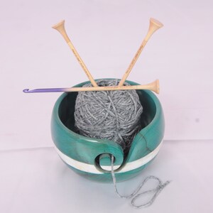 May include: A teal wooden yarn bowl with a white stripe, holding a ball of gray yarn. Two wooden knitting needles and a purple crochet hook are resting on top of the yarn.