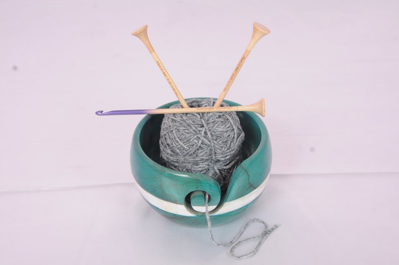 May include: A teal wooden yarn bowl with a white stripe, holding a ball of gray yarn. Two wooden knitting needles and a purple crochet hook are resting on top of the yarn.
