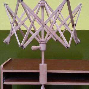 May include: A wooden yarn swift with a crank handle, mounted on a wooden table with a drawer. The swift is in a starburst shape with multiple arms.