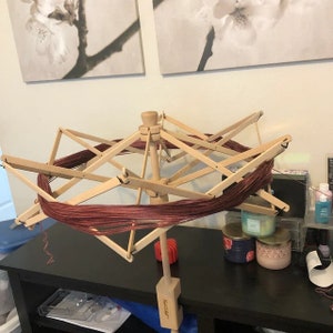 May include: A wooden yarn swift with a red yarn wound around it. The swift is on a black table with candles and other items.