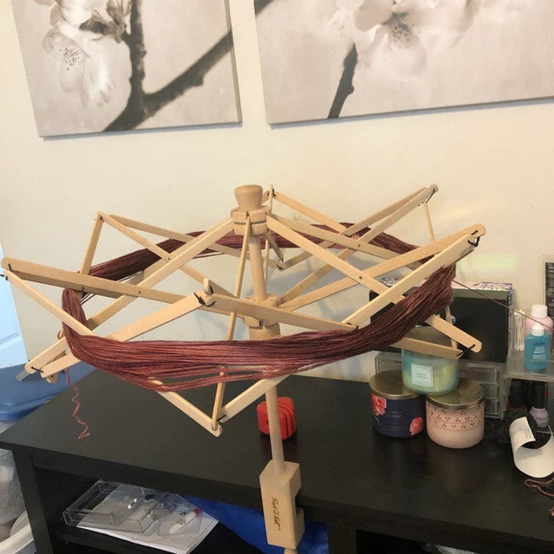 May include: A wooden yarn swift with a red yarn wound around it. The swift is on a black table with candles and other items.