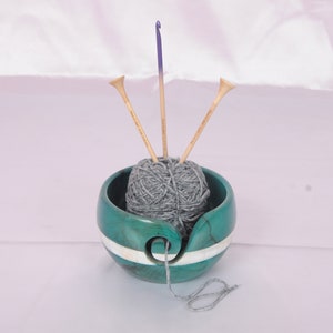 May include: A teal yarn bowl with a white stripe, holding a ball of gray yarn and three knitting needles.