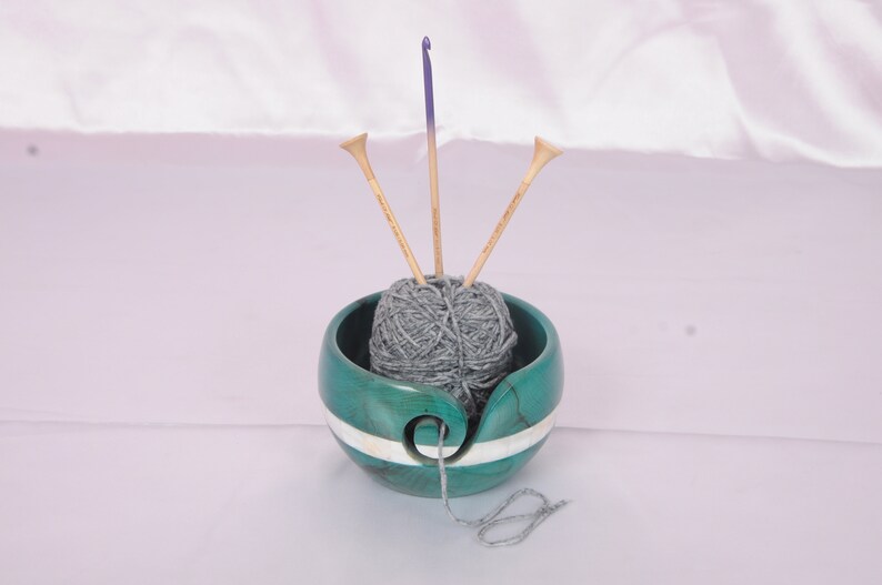 May include: A teal yarn bowl with a white stripe, holding a ball of gray yarn and three knitting needles.