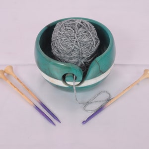 May include: A teal wooden yarn bowl with a white stripe and a gray yarn ball inside. Two purple and natural wood knitting needles are on a white surface.