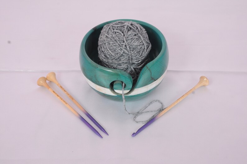May include: A teal wooden yarn bowl with a white stripe and a gray yarn ball inside. Two purple and natural wood knitting needles are on a white surface.