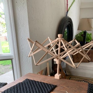 May include: A wooden yarn swift with a rotating spindle and multiple arms for winding yarn. The swift is on a wooden table with a black quilted placemat.