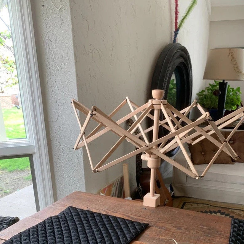 May include: A wooden yarn swift with a rotating spindle and multiple arms for winding yarn. The swift is on a wooden table with a black quilted placemat.