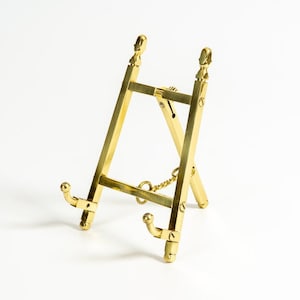 May include: A gold-toned metal easel stand with a decorative design. The stand is made of two pieces that fold together for easy storage.