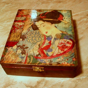 Pode incluir: Uma caixa de madeira com tampa articulada, com uma ilustração de gueixa japonesa. A obra de arte na tampa mostra uma gueixa em um quimono colorido com padrões florais, em um fundo azul claro. A caixa tem uma base marrom escuro e um fecho dourado.