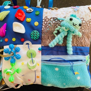 Peut inclure: Un coussin sensoriel multicolore avec différentes textures et objets attachés. Le coussin est en tissu bleu, blanc et vert avec une poche zippée bleue. Il y a différents boutons, une petite pieuvre bleue en crochet et un jouet en plastique vert attachés au coussin.