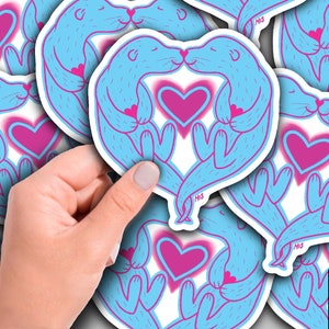 May include: A set of blue and pink otter stickers with hearts. The otters are holding hands and kissing. The stickers are on a white background.