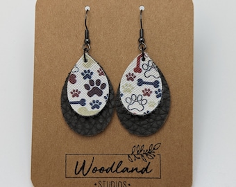 Puede incluir: Un par de pendientes colgantes con una base de cuero negro y una parte superior de tela blanca, roja y azul con huellas de patas y diseños de huesos. Los pendientes se muestran en una tarjeta marrón con el texto "Woodland Studios".