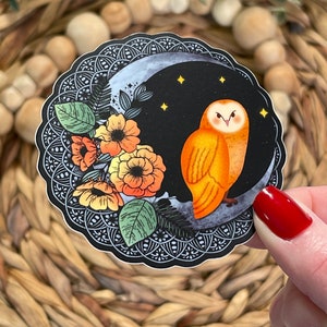 Pu&ograve; includere: Un adesivo con un'illustrazione stilizzata di un gufo arancione appollaiato su una luna crescente con stelle. Il gufo &egrave; circondato da fiori stilizzati in tonalit&agrave; di arancione, giallo e rosso. Lo sfondo &egrave; nero con un bordo decorativo.