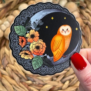 Pu&ograve; includere: Un adesivo con un'illustrazione stilizzata di un gufo arancione appollaiato su una luna crescente con un design floreale. Il gufo ha grandi occhi rotondi e un piumaggio dettagliato. Lo sfondo &egrave; nero con stelle bianche.
