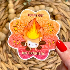 May include: A colorful sticker featuring a marshmallow character on a campfire. The marshmallow is on fire, with the text "BURNT OUT" above and "BUT OPTIMISTIC" below. The sticker has a floral design with orange, pink, and yellow hues.