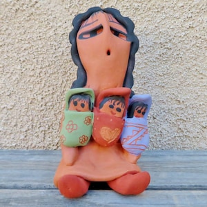 May include: A terracotta figurine of a woman with a long, dark brown hair, holding three babies wrapped in colorful blankets. The woman has a large, round face with exaggerated features and a wide, open mouth. The babies are all smiling and have dark hair and eyes.