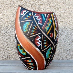 May include: A hand-painted ceramic vase with a black background and colorful geometric patterns. The vase features a vertical orange stripe and a curved shape. The design includes red, white, yellow, and turquoise accents.
