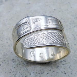 May include: A silver ring with intricate engraved designs. The ring features a wrap-around style with geometric and stylized patterns. The interior of the ring is stamped with the letters "R C".