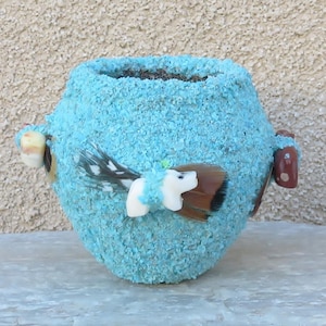 May include: A turquoise-colored, textured ceramic vase with decorative elements. The vase features feather and white animal-shaped embellishments, along with brown and tan accents. The vase is designed for decorative purposes.