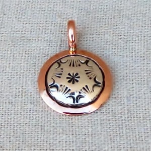 May include: A circular pendant featuring a polished copper outer ring and a central silver-toned disc. The central disc is decorated with a stamped star-like pattern surrounded by radiating lines and curved motifs, topped with a small copper bail.