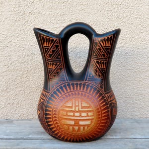 May include: A black and orange ceramic vessel featuring a central circular motif with geometric patterns. The vase has a distinct bridge handle connecting two flared openings, with intricate etched lines covering the entire surface.