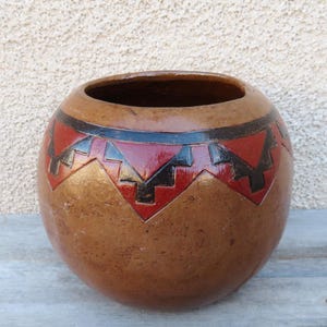 May include: A round, brown ceramic pot with a wide opening. The pot features a decorative band of red and black geometric patterns around the top. The pot has a textured surface and is likely handmade.