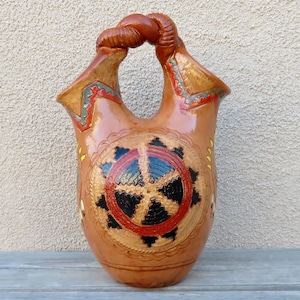 Puede incluir: Un jarrón de cerámica marrón con un diseño de asa único. El jarrón presenta intrincados patrones en rojo, negro y amarillo, con un diseño circular central. El asa es un diseño de cuerda trenzada.