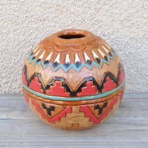 May include: A round, brown, hand-carved decorative vessel with a small opening at the top. The vessel features intricate geometric patterns in red, black, white, and turquoise. The design includes zigzags and stepped shapes, creating a Southwestern aesthetic.