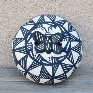 May include: A round, white ceramic decorative piece with a black geometric pattern and a black butterfly accent. The butterfly has yellow details. The piece is sitting on a wooden surface.
