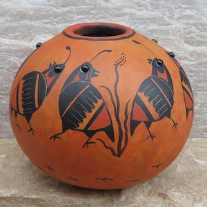 Op de afbeelding: Een handgeschilderde kalebasvaas met drie zwarte en oranje vogels, elk met een zwart oog, op een oranje achtergrond. De vaas heeft een bruine rand en zwarte stippen rond de bovenkant.