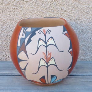 May include: A round, hand-painted ceramic pot with a wide opening. The pot is primarily orange with a cream-colored central design featuring stylized floral motifs in black, gray, and red. The pot sits on a wooden surface.