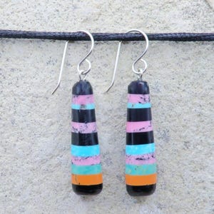 May include: A pair of drop earrings with silver hooks. Each earring has a black, pink, turquoise, and orange striped design. The earrings are suspended against a textured, light grey background.