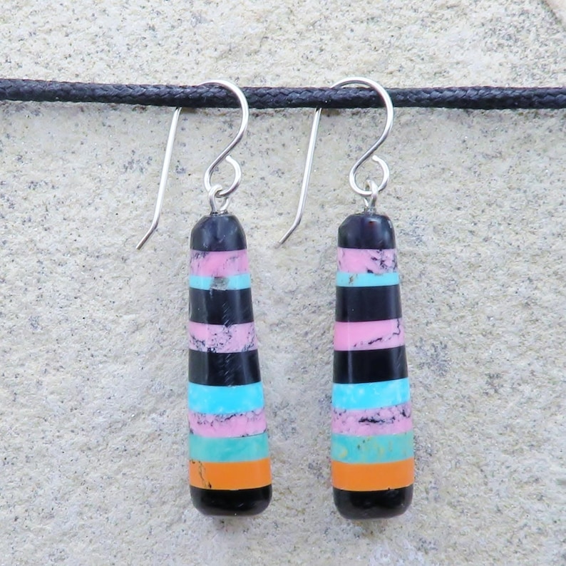 May include: A pair of drop earrings with silver hooks. Each earring has a black, pink, turquoise, and orange striped design. The earrings are suspended against a textured, light grey background.