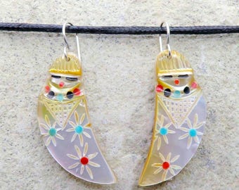 May include: A pair of handcrafted earrings with a distinctive design. Each earring is crescent-shaped, depicting a figure with a hat and colourful accents. The earrings have a light, pearlescent finish and are suspended from silver-tone hooks.