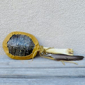 May include: A rattle constructed from a dark, patterned turtle shell encased in a mustard yellow leather border. The handle is a light-colored bone, and the base of the handle is adorned with a single dark grey feather tied with thin leather cords.