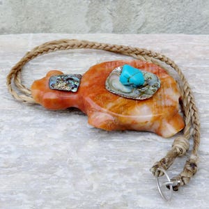 May include: A carved orange stone turtle pendant with turquoise and abalone shell accents. The pendant is on a braided leather cord.