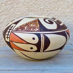 May include: A white ceramic pot with a geometric design in brown, orange, and black. The pot has a rounded shape and a wide opening.