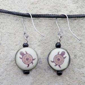 May include: A pair of dangle earrings featuring round, off-white beads with a brown turtle design. The earrings have silver-colored hooks and black beads. The earrings are hanging from a black cord.