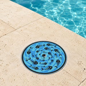 May include: A circular pool drain cover featuring a blue mosaic-style background with a repeating pattern of black fish skeletons arranged in a spiral design, centered around a small circular opening in the middle of the disc.