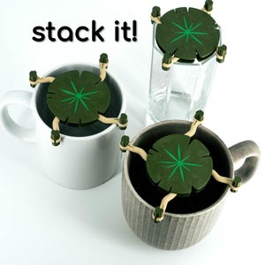 May include: Three green and brown leaf-shaped coasters with a star design in the center. The coasters are stacked on top of each other. The text "stack it!" is written in black letters on a white background.